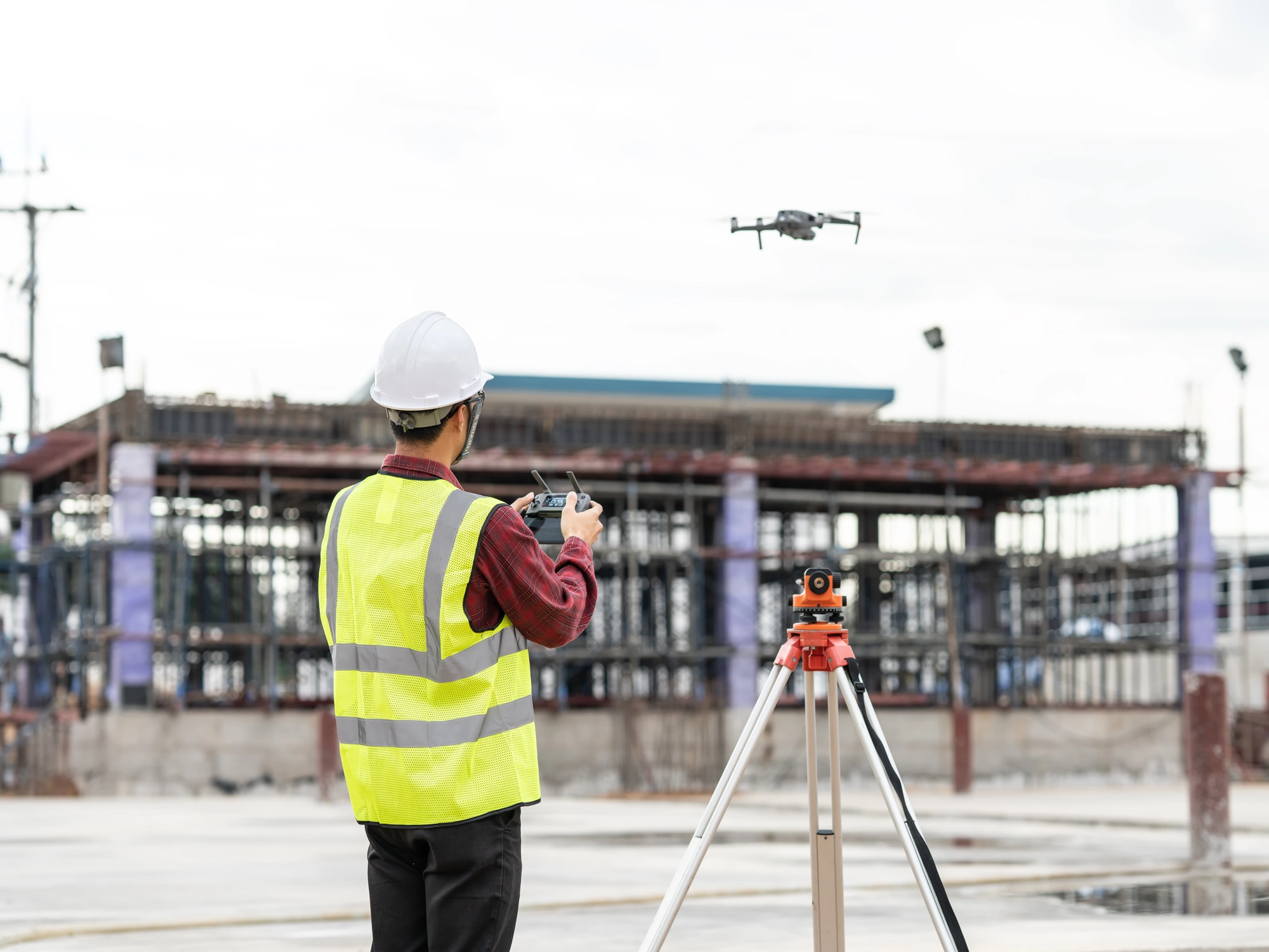 Man in hi-vis vest and white hard hat uses total station and drone to gather valuable survey data on construction site.