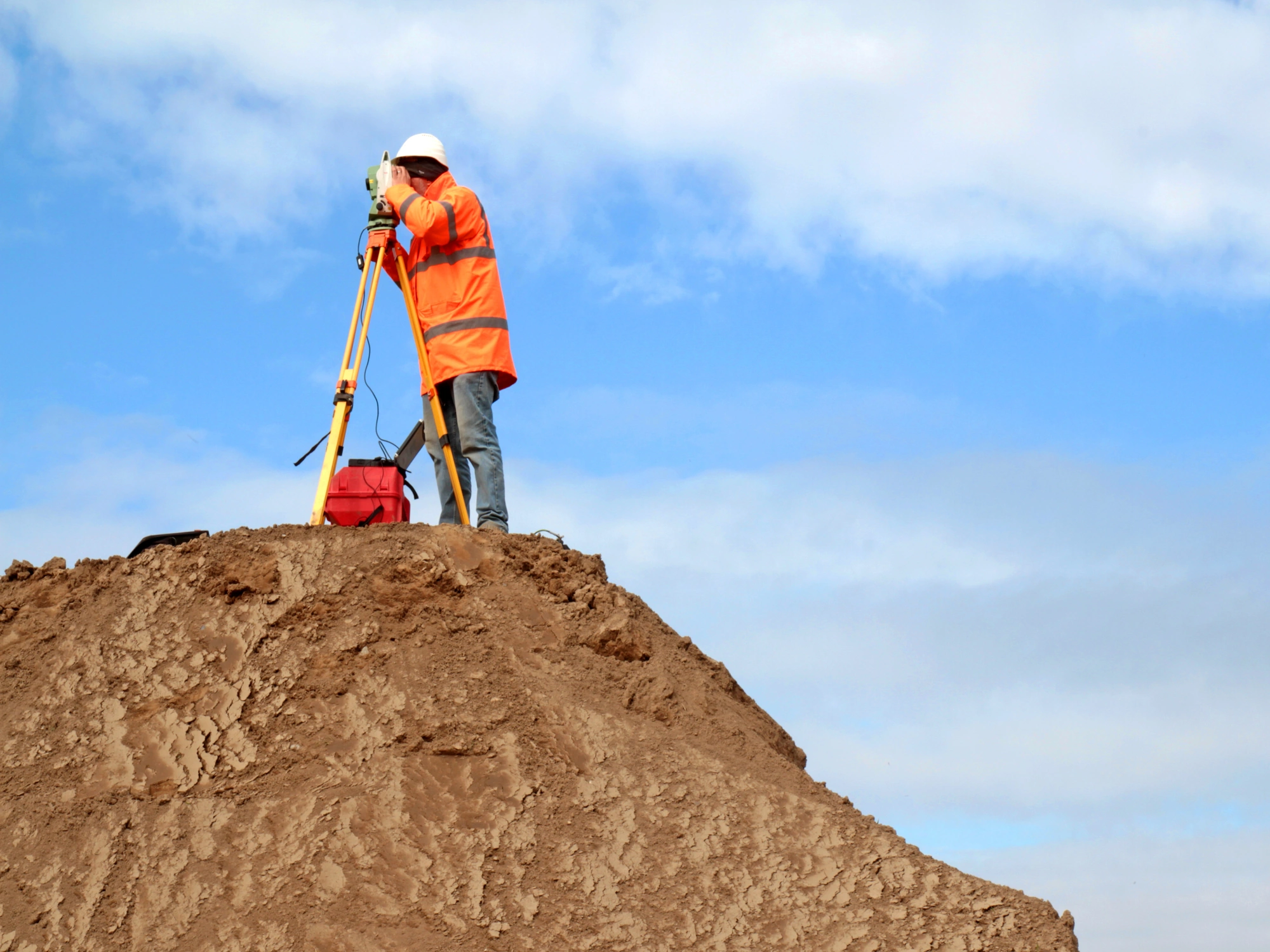 A surveyor in a white hardhat and orange jacket uses surveying equipment on top of a large dirt mound.