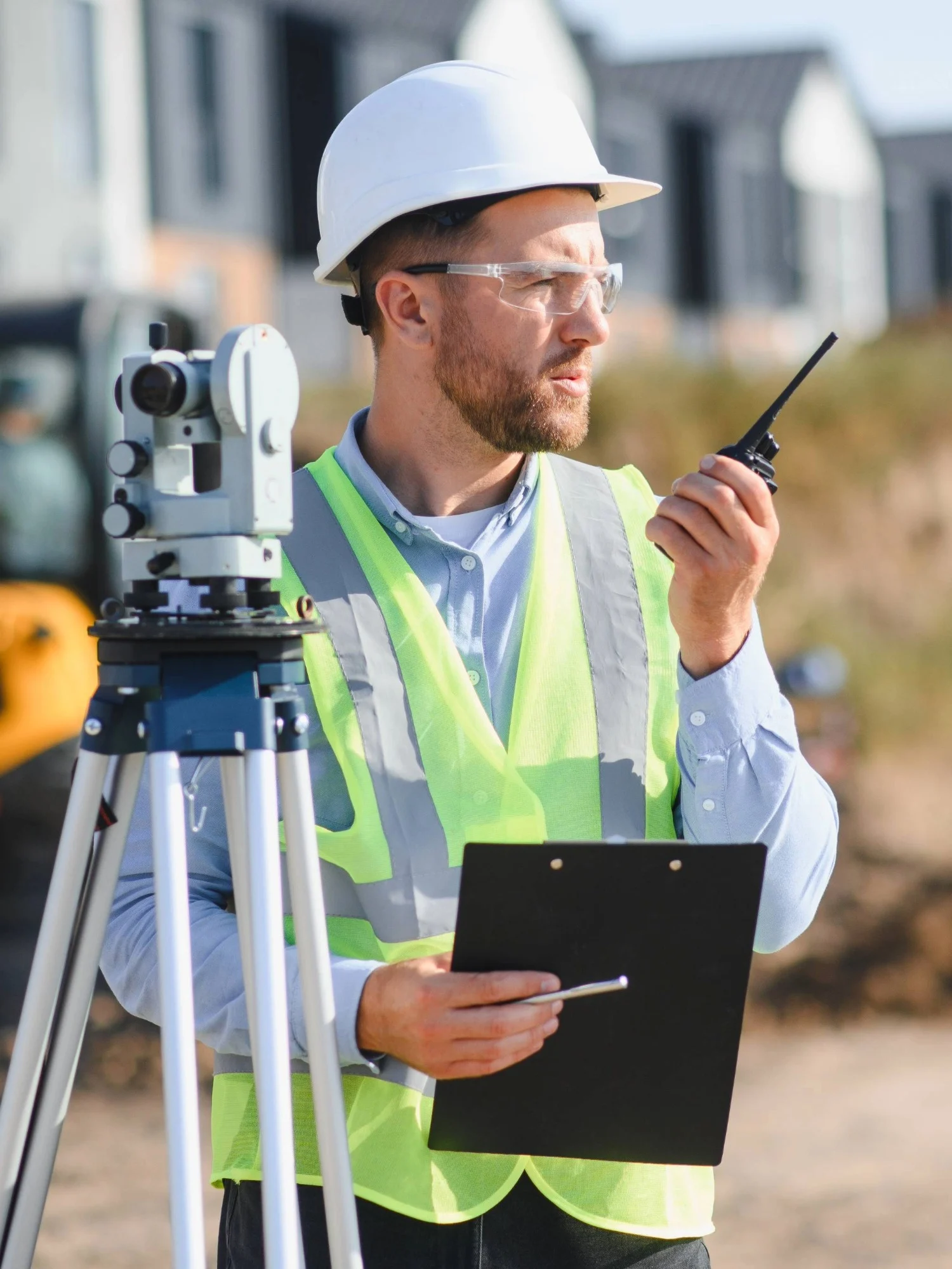 A man in hi-vis jacket and hard hat stands next to survey equipment on a construction site.