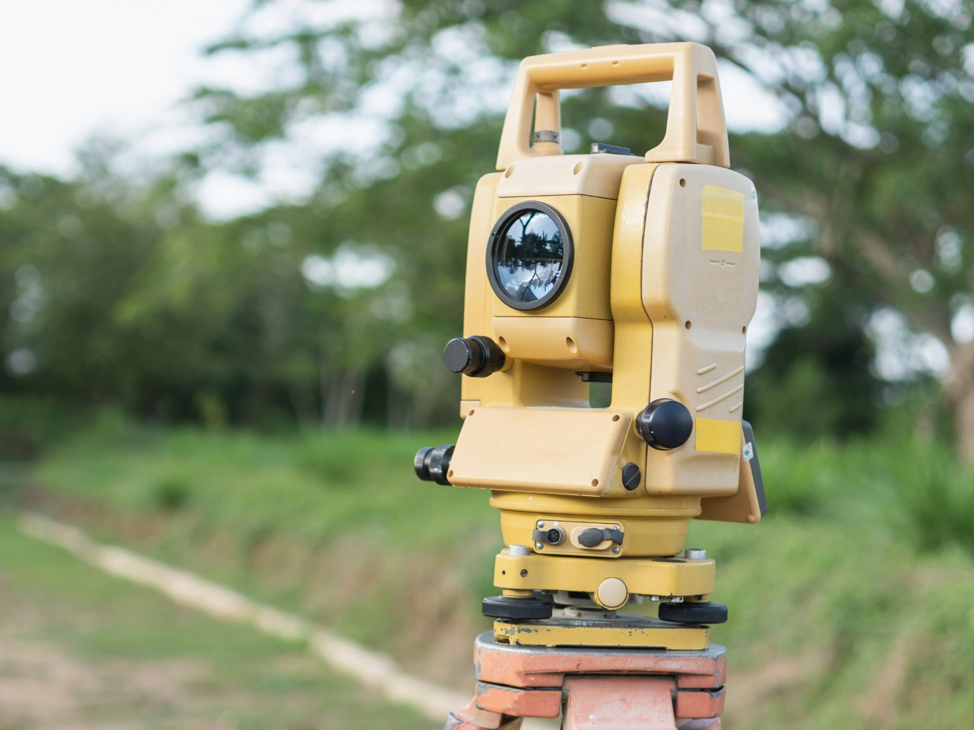 A yellow total station on a field with a trodden path and trees behind it.