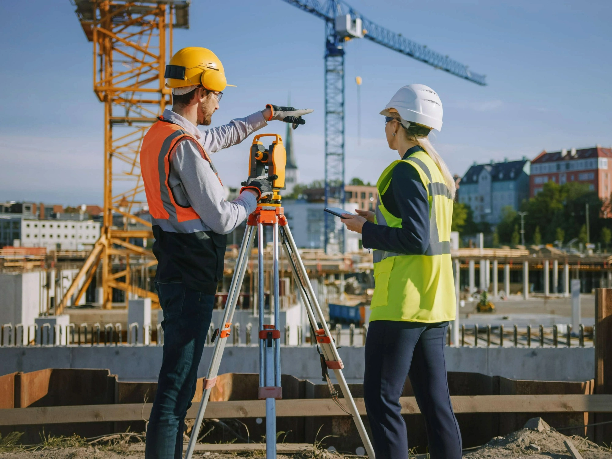 Land surveyors on a construction site, using a total station during their survey with cranes in background.