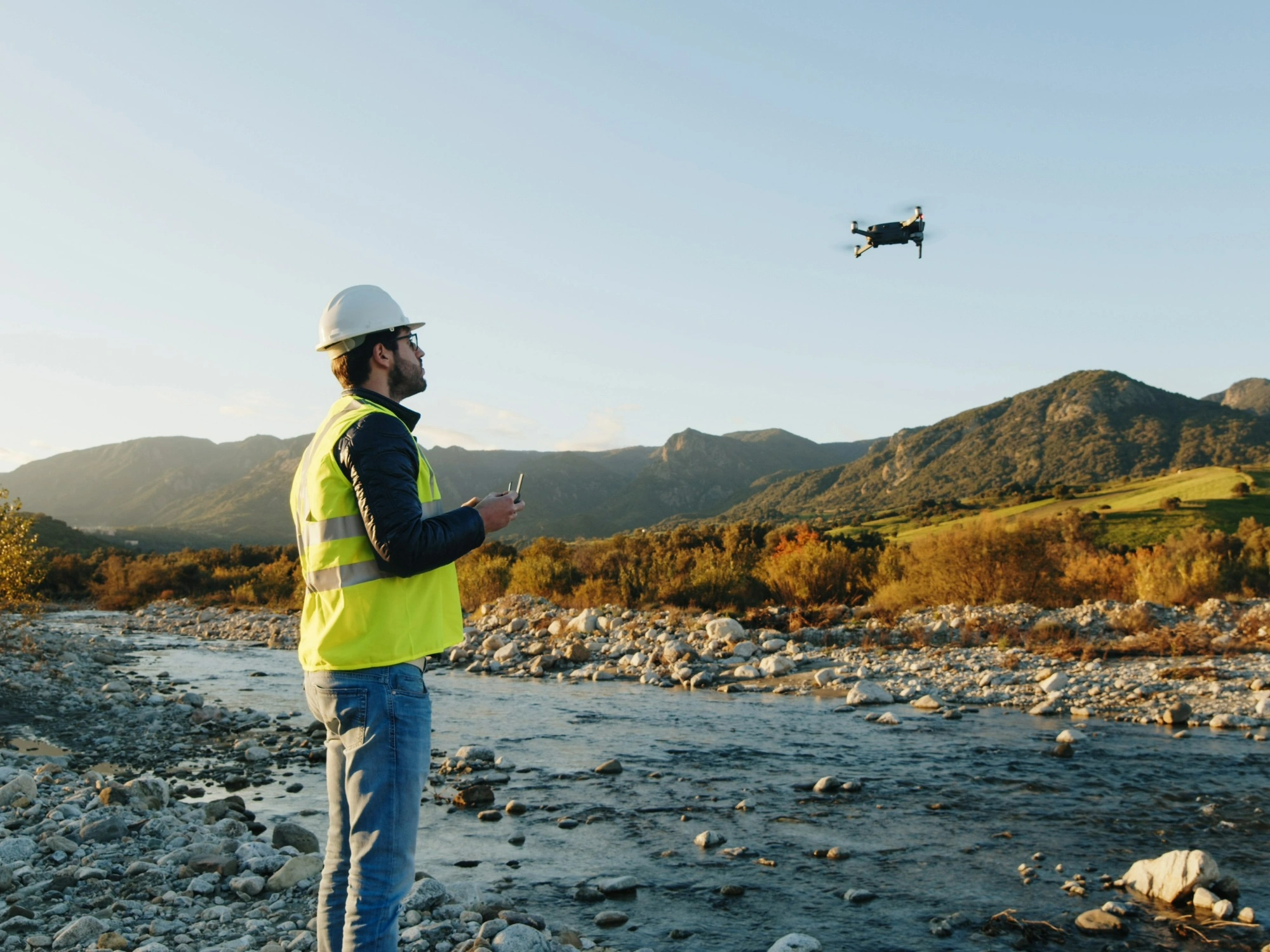 Land surveyor flying drone over environmental area with a shallow river and hills.